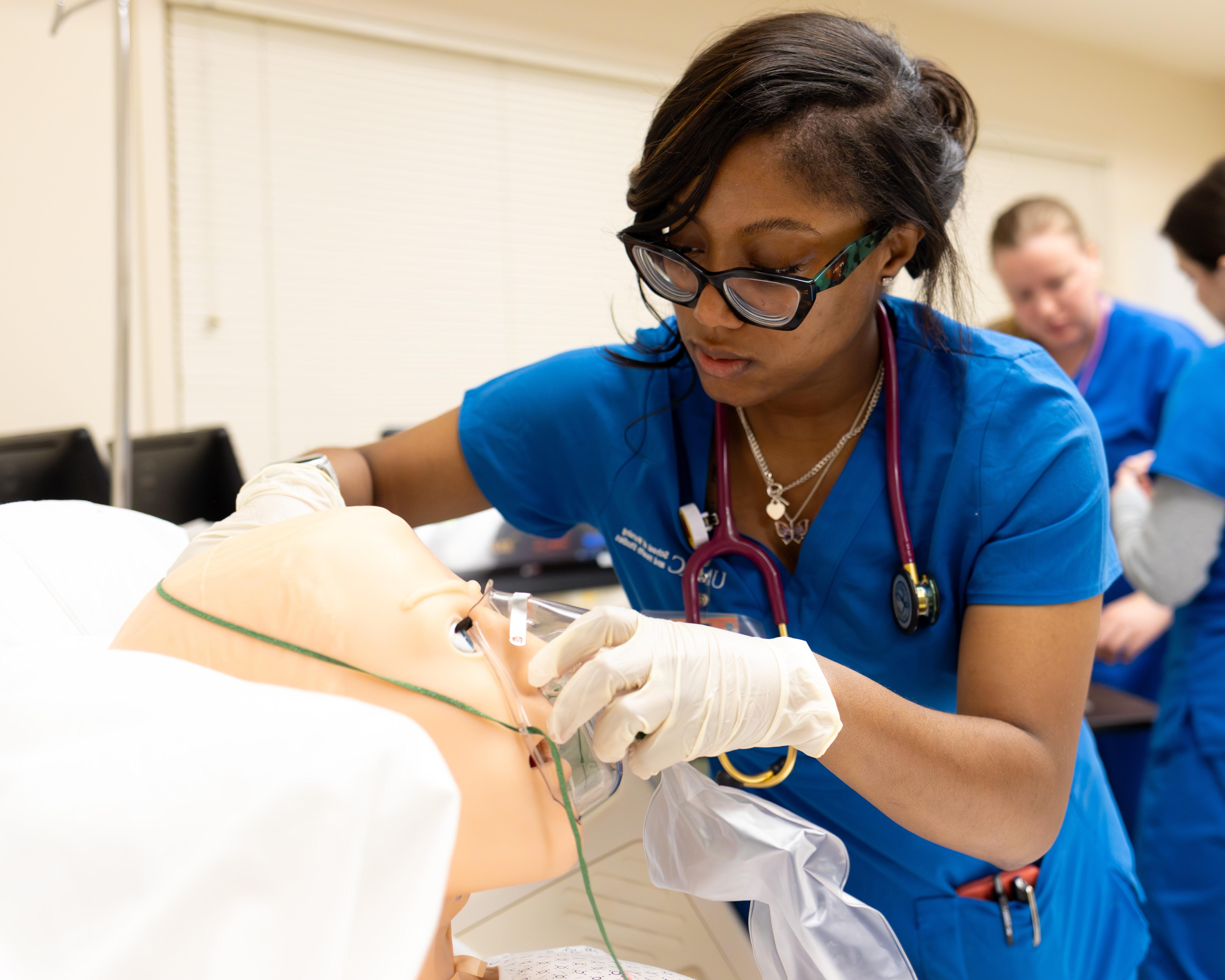 Nursing student Tatyana Charles wearing blue nursing scrubs with a stethoscope around her neck places an oxygen mask on a dummy in the nursing simulation lab.